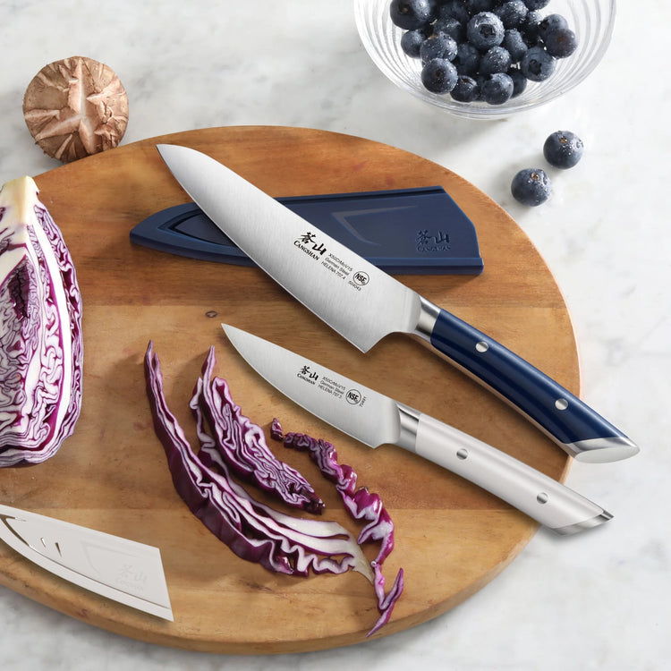 Two knives with navy and white handles on a wooden board, one with a sheath, next to sliced purple cabbage and blueberries.