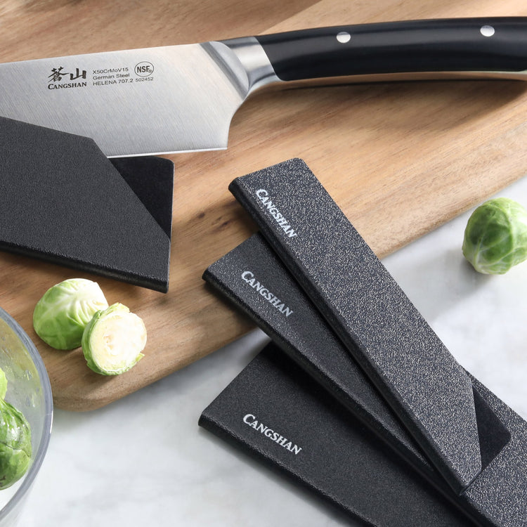 Set of six black knife edge guards with textured surfaces on a wooden cutting board next to a kitchen knife and Brussels spro