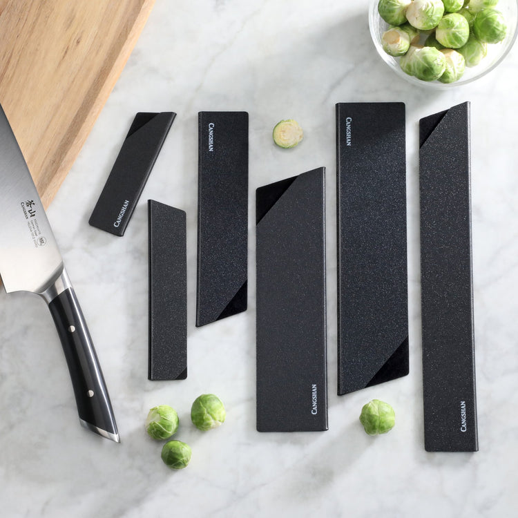 Six black knife edge guards of varying lengths arranged on a marble surface next to a chef's knife and Brussels sprouts.