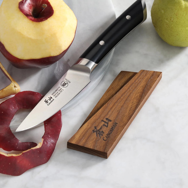 Paring knife with black handle next to peeled apple and ash wood magnetic knife sheath on marble surface.