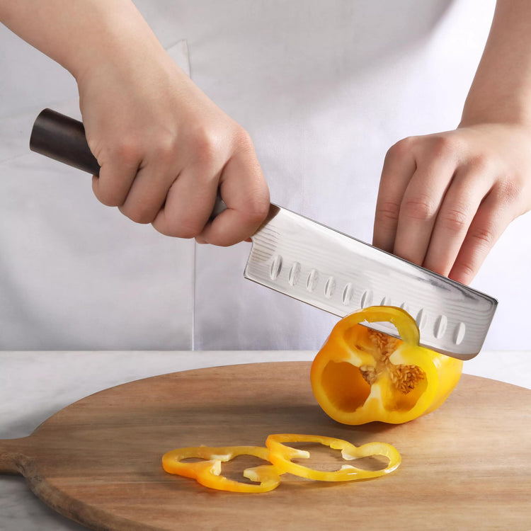 Hands slicing a yellow bell pepper on a wooden cutting board with a Nakiri knife.