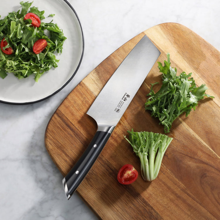 7-inch Nakiri knife with black handle on wooden cutting board next to chopped greens and halved cherry tomato.