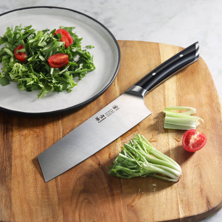 7-inch Nakiri knife with black handle on wooden cutting board next to chopped celery and tomato, plate with salad nearby.
