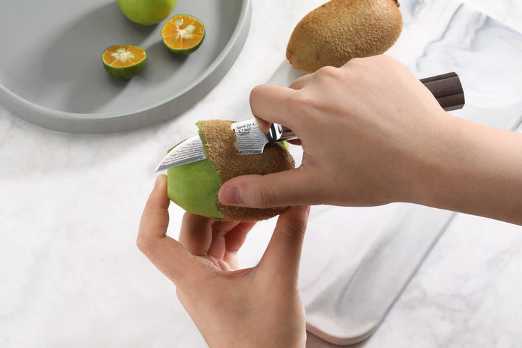 Hands using the MAYA Series 2.75-inch Damascus steel peeling knife to peel a kiwi.
