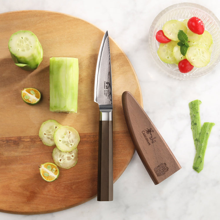 MAYA Series 3.5-Inch Damascus Steel Paring Knife with wooden sheath on a cutting board next to sliced cucumbers.