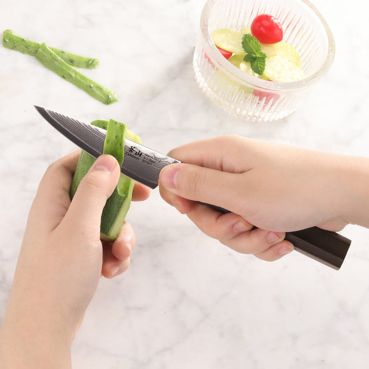 Hands peeling a cucumber with the MAYA Series 3.5-inch paring knife in X-7 Damascus steel.