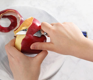 
                    
                      Hands peeling a red apple with a small blue-handled knife on a white plate.
                    
                  