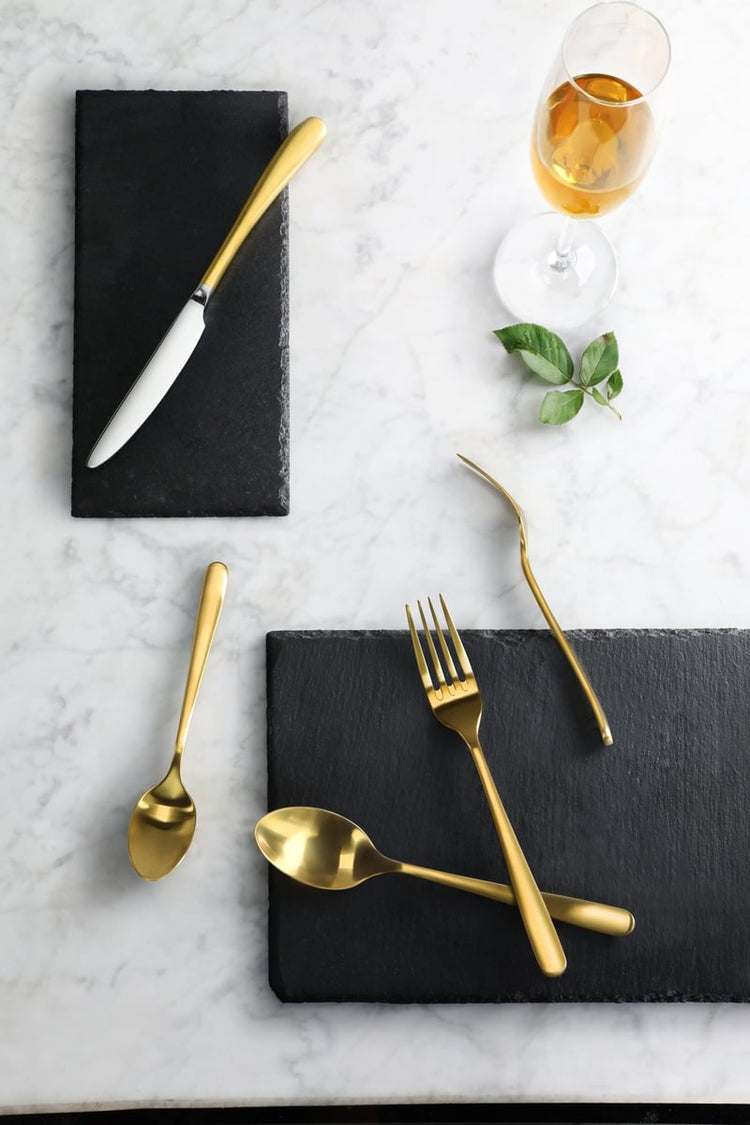 Gold RAIN II Series flatware set displayed on dark slate tiles over a marble surface.