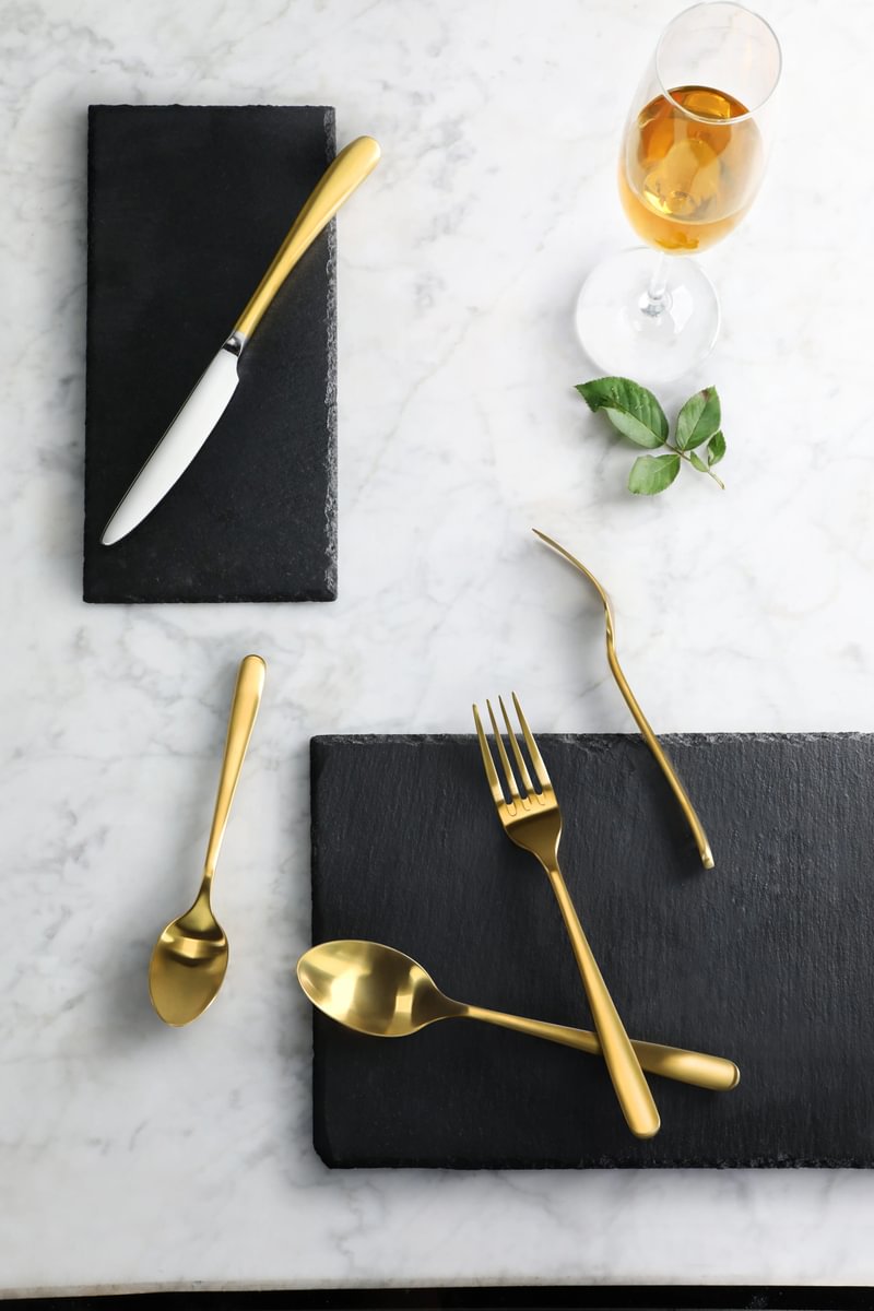 Brushed gold flatware set with knife, fork, spoons on black slate plates and a glass of amber liquid on marble surface.