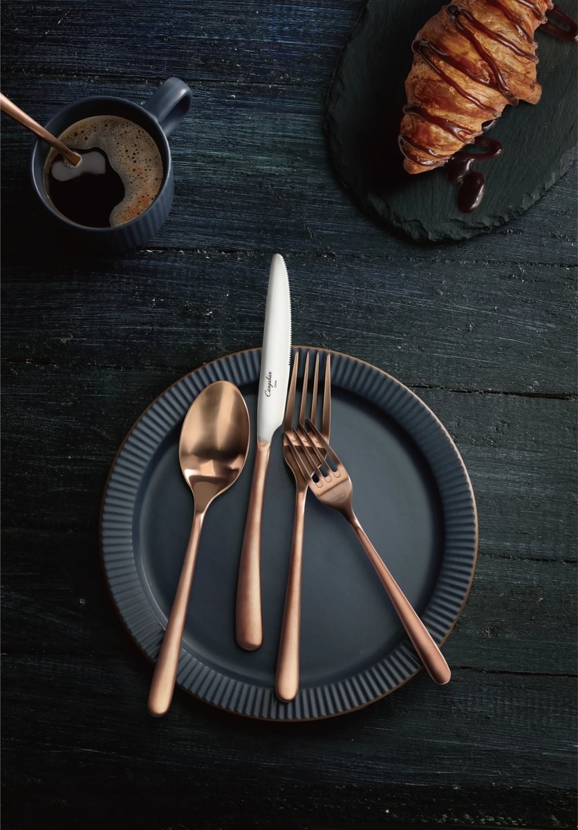 Brushed copper spoon, knife, and fork on a dark plate with a croissant drizzled in chocolate and a cup of coffee.