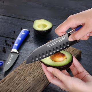
                    
                      Hands holding a halved avocado, using a knife to remove the pit on a wooden surface with another knife nearby.
                    
                  