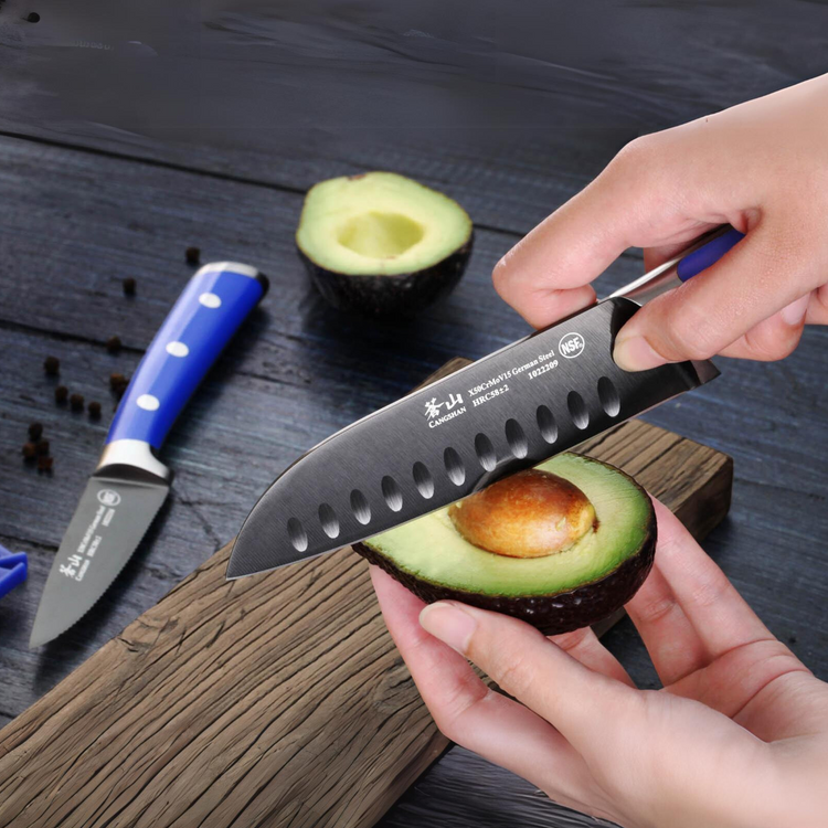Hands holding a halved avocado, using a knife to remove the pit on a wooden surface with another knife nearby.
