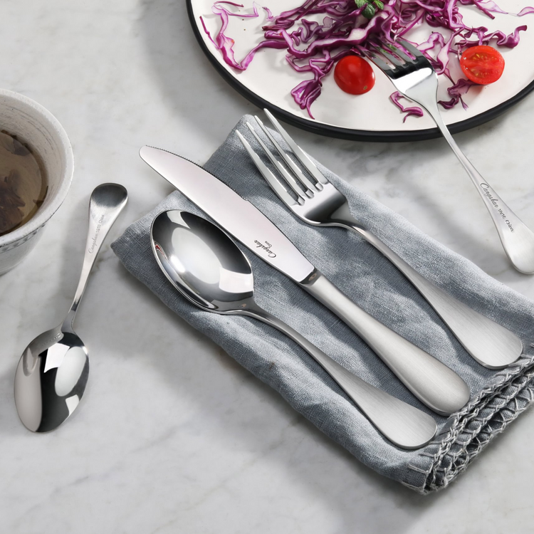Stainless steel flatware set with matte finish handles, including knife, fork, spoon, and teaspoon on a gray cloth.