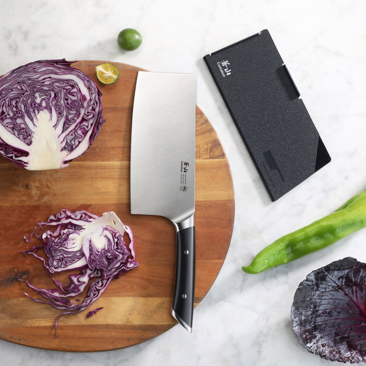 7-inch vegetable cleaver with black handle on wooden board, sliced purple cabbage, lime, and black knife sheath nearby.