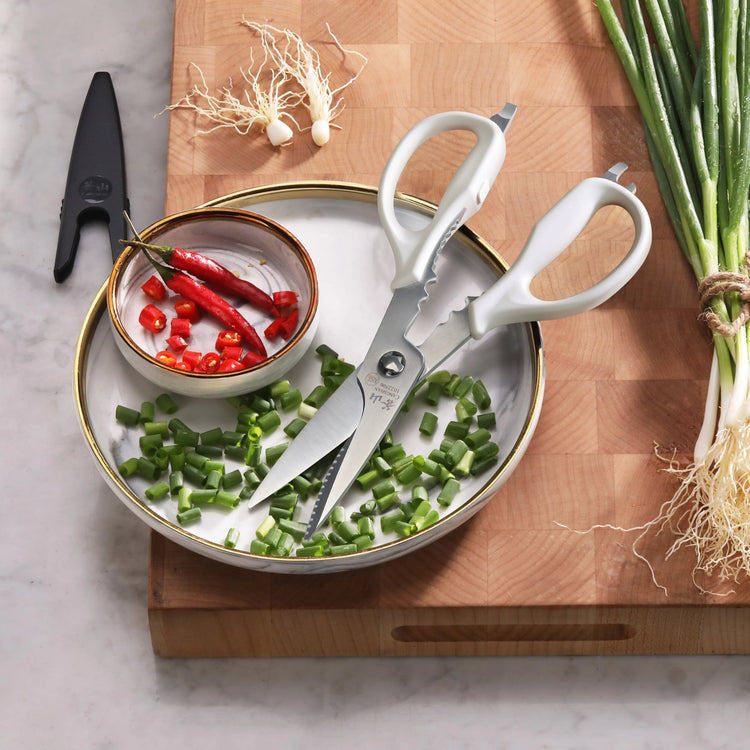 White-handled kitchen shears with blade holder beside chopped green onions and red chili peppers on a plate.