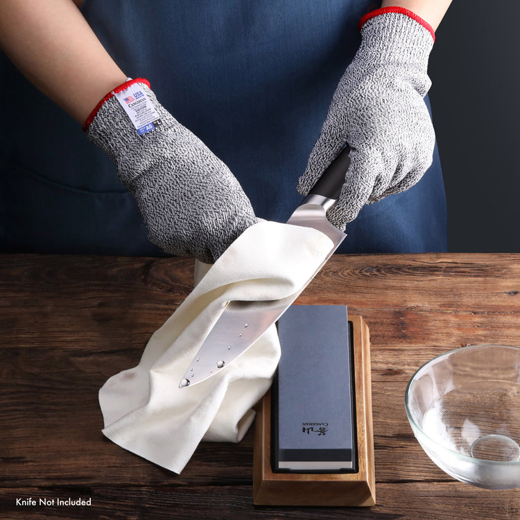 Hands wearing cut-resistant gloves holding a knife being wiped with a cloth next to a sharpening stone and water bowl.