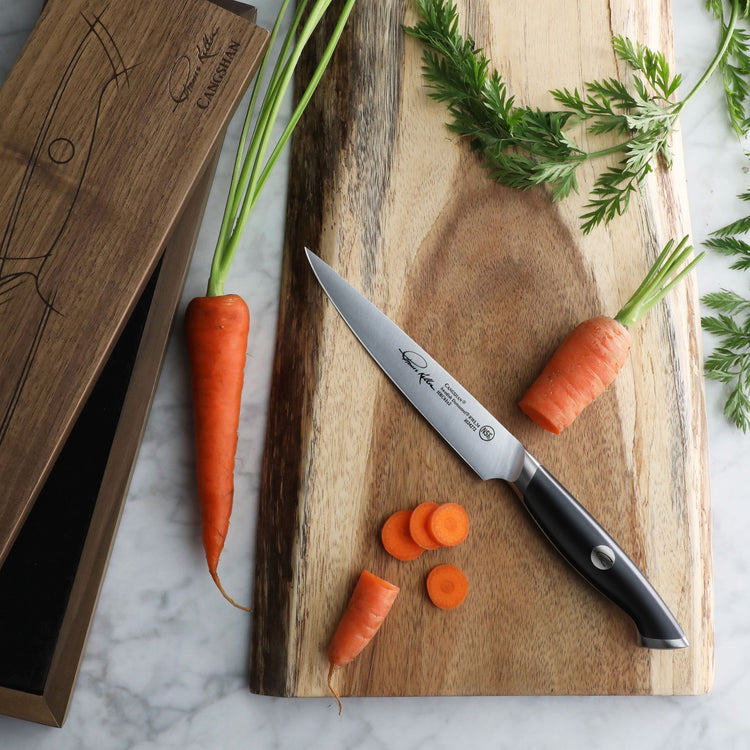 Utility knife with black handle on wooden cutting board next to whole and sliced carrots and wooden box.