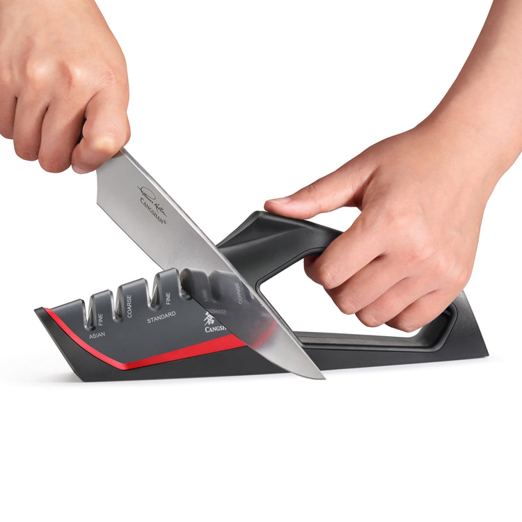 Hands using a red and black handheld knife sharpener with coarse, standard, and ceramic slots to sharpen a kitchen knife.