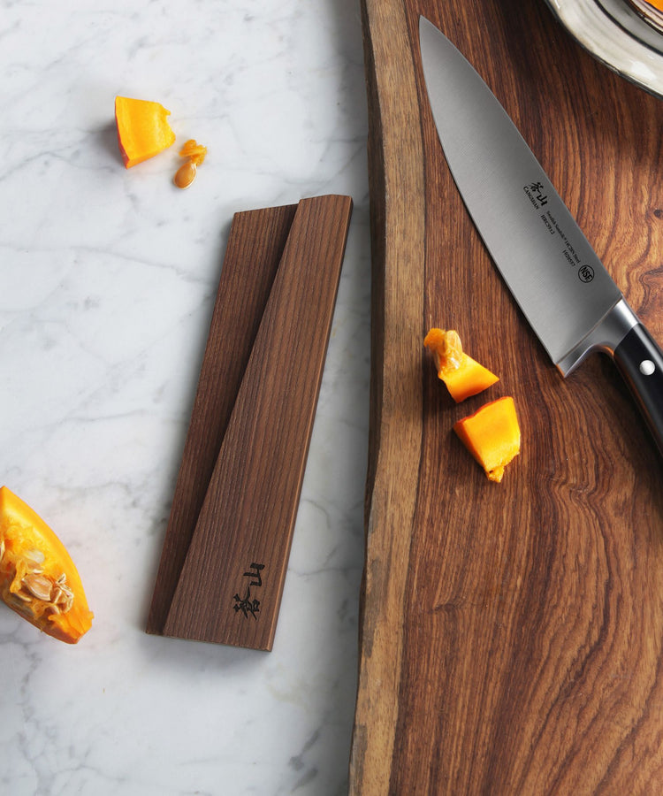 Solid ash wood magnetic knife sheath placed on a marble surface next to a wooden cutting board with a chef's knife and choppe
