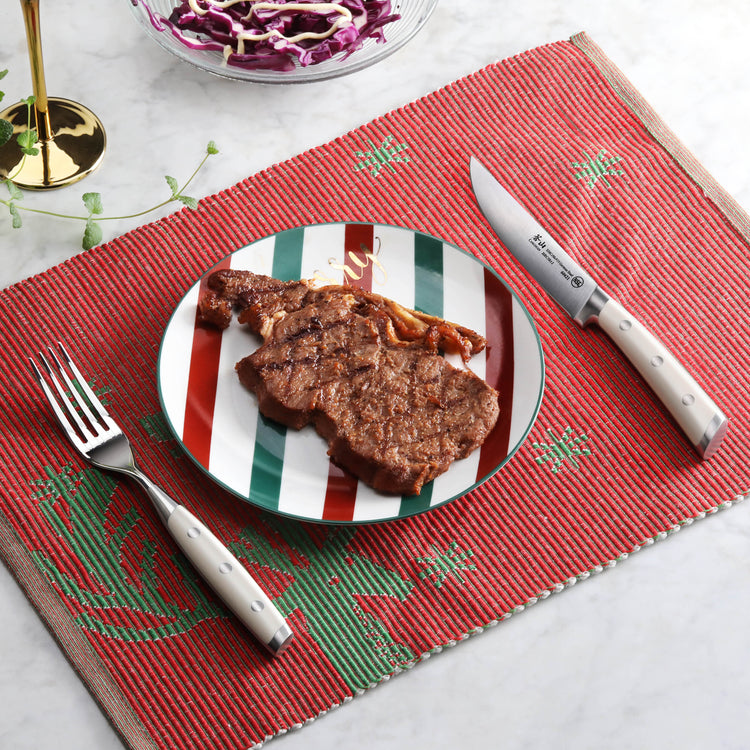Grilled steak on striped plate with steak fork and serrated knife on red placemat.