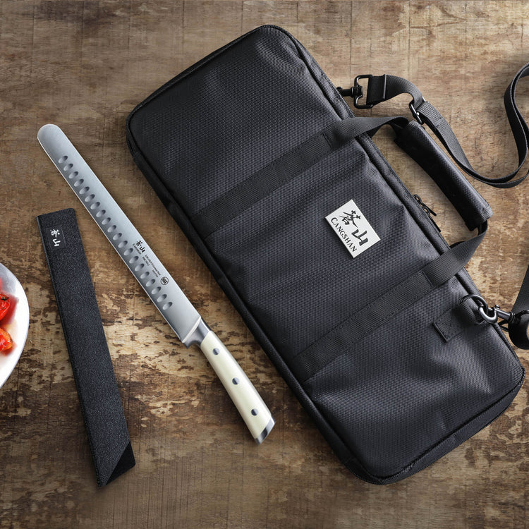 Black nylon knife bag with strap and handles, lying flat on a wooden surface next to a long knife with a white handle and bla