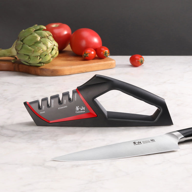 Black and red handheld knife sharpener with coarse and fine slots next to a chef's knife on a marble surface.