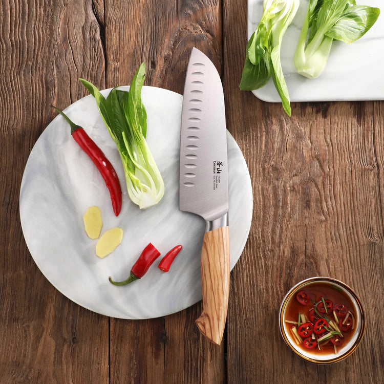 Santoku knife with wooden handle on a round marble board with chili peppers, bok choy, and ginger slices.