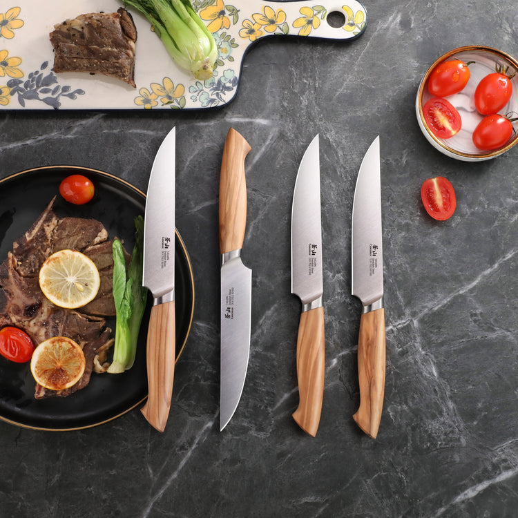 Four steak knives with olivewood handles and stainless steel blades arranged on a dark surface next to plates with steak, lem