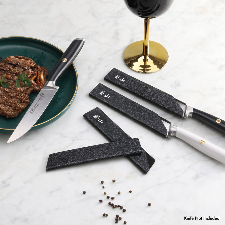 Three steak knives with black textured guards on the blades, placed next to a cooked steak on a green plate and a glass of re