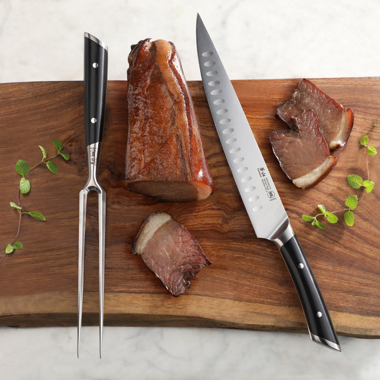 Carving fork and knife with black handles on a wooden board next to sliced cooked meat and herbs.