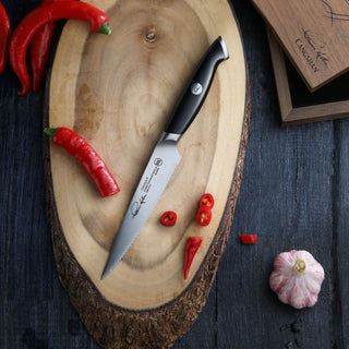 
                    
                      5-inch serrated utility knife with black handle on wooden cutting board, surrounded by red chili peppers and garlic bulb.
                    
                  