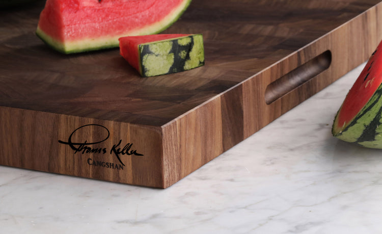 Walnut end-grain cutting board with engraved Thomas Keller signature and watermelon slices on top and beside it.