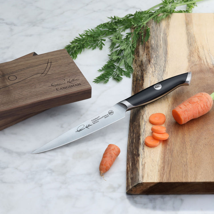 Black-handled fine-edge utility knife with engraved blade, placed on a wooden cutting board next to sliced carrots and a wood