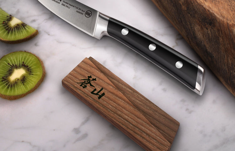 Solid ash wood magnetic knife sheath with black Japanese characters, placed next to a chef's knife and sliced kiwi on a marbl