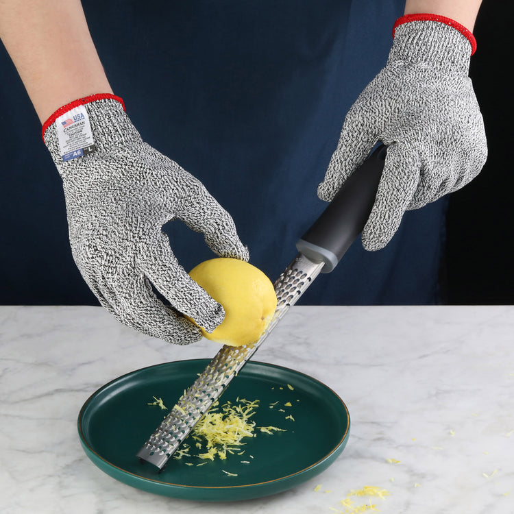 Hands wearing gray cut resistant gloves grating a lemon over a green plate on a marble surface.