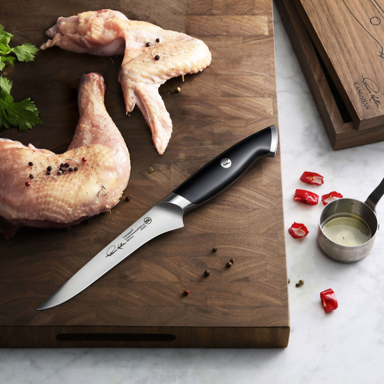 Boning knife with black handle on wooden cutting board next to raw chicken wings and scattered peppercorns.