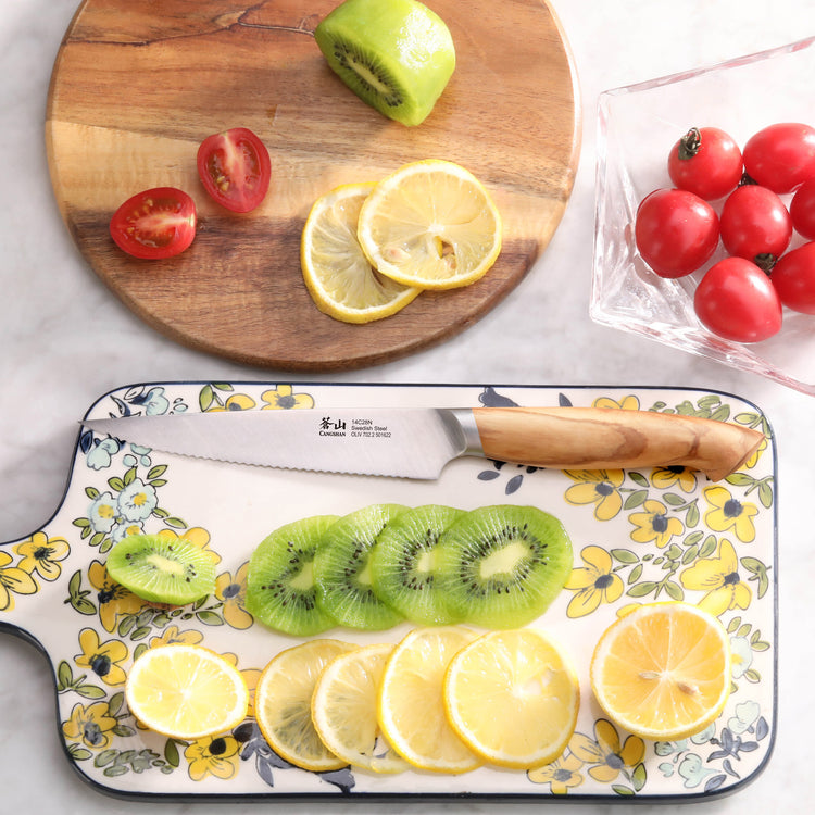 Serrated utility knife with olivewood handle on floral cutting board, sliced kiwi and lemon, halved cherry tomatoes on wooden