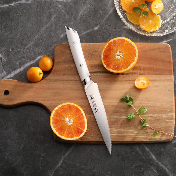 Serrated utility knife with white handle on wooden cutting board next to sliced oranges and small tomatoes.