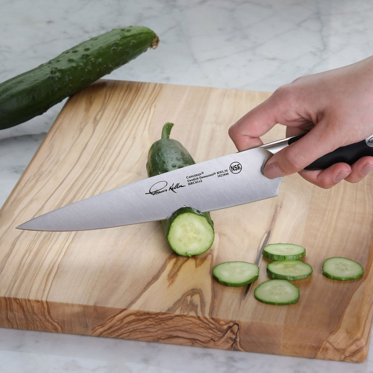 Hand holding a utility knife slicing a cucumber on a wooden cutting board with cucumber slices nearby.