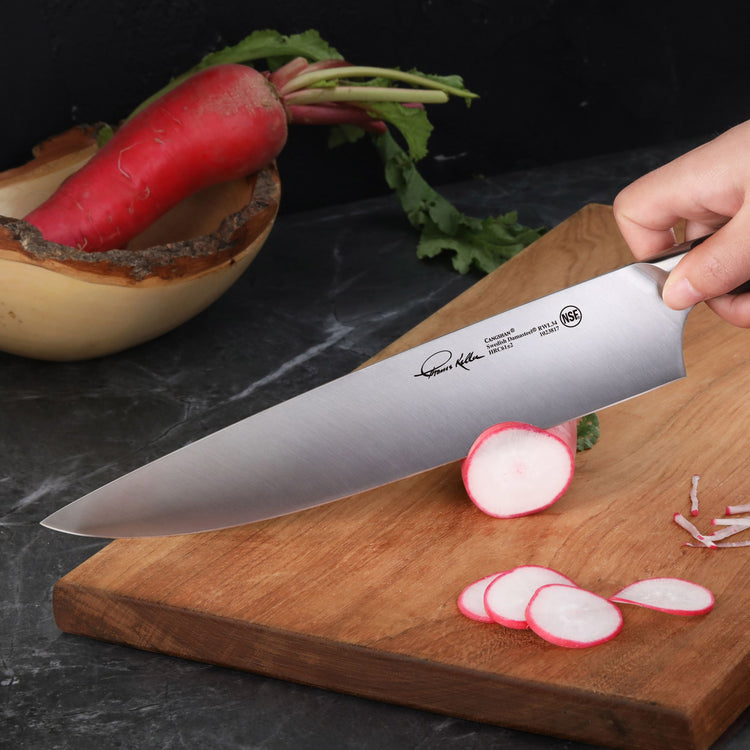 Hand holding a chef's knife slicing radishes on a wooden cutting board with whole radishes in a bowl nearby.