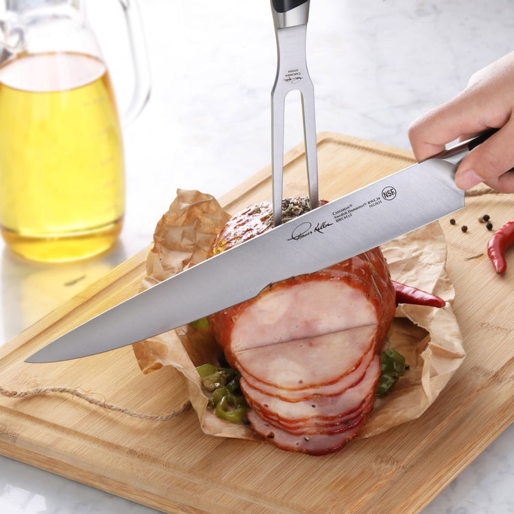Hand holding a carving knife slicing a cooked ham secured with a fork on a wooden cutting board.