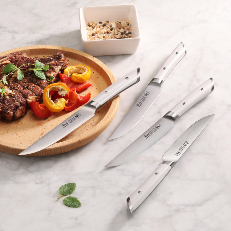 Four stainless steel steak knives with white handles arranged on a marble surface next to a wooden plate with grilled steak a