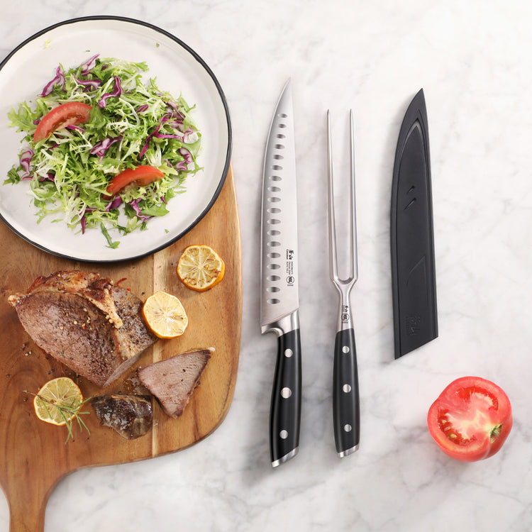 Two-piece carving set with black handles, including a serrated knife and carving fork, next to a sheath, sliced meat, salad,