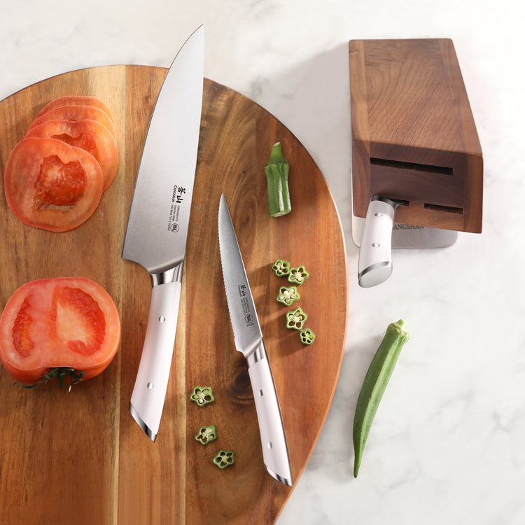 Two stainless steel knives with white handles on a wooden cutting board with sliced tomato and okra, next to a wooden knife b