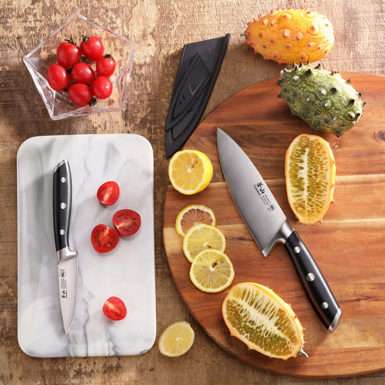 Two black-handled knives with sheaths, one cutting cherry tomatoes on a marble board, the other slicing exotic fruits on a wo