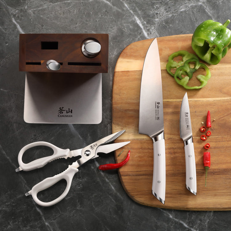 Two kitchen knives with white handles on a wooden cutting board next to sliced green bell pepper and red chili peppers, white