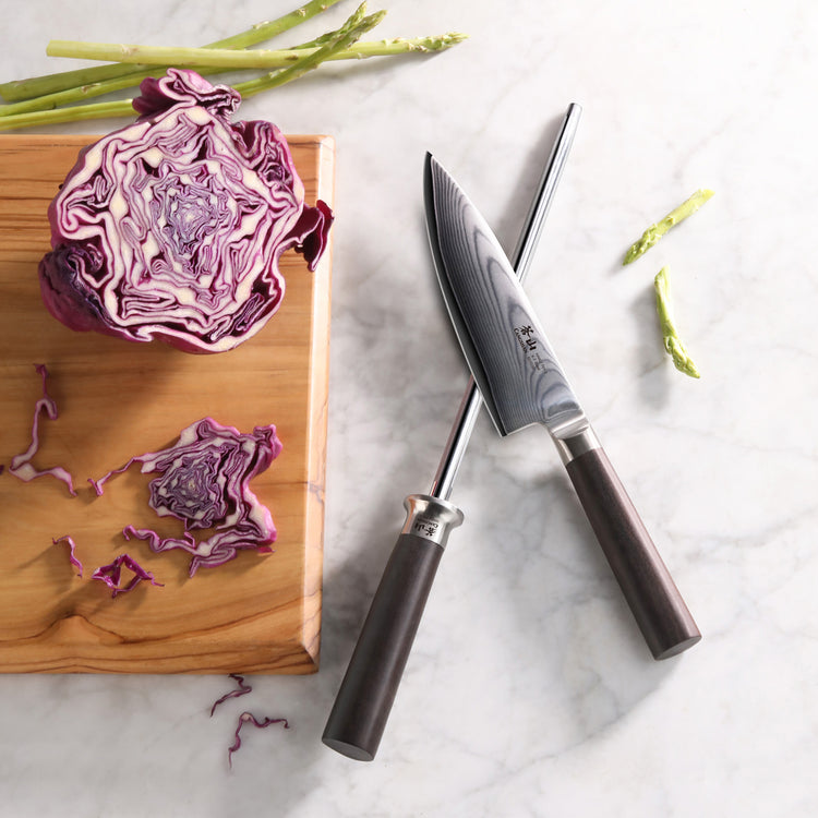 Crossed kitchen knife and honing steel with dark handles on marble surface next to chopped purple cabbage on wooden board.