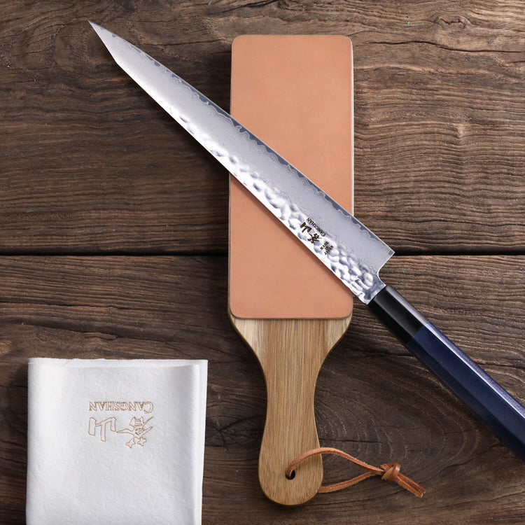 Japanese-style knife resting on a 2-sided leather paddle strop with a wooden handle and a white cloth nearby.