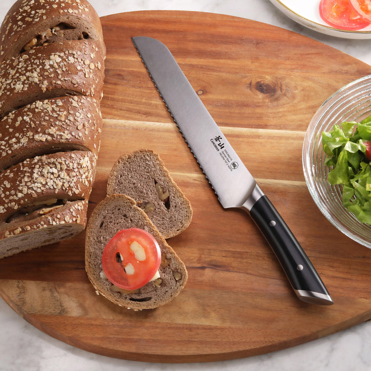 Bread knife with black handle on wooden cutting board next to sliced whole grain bread and tomato slice.