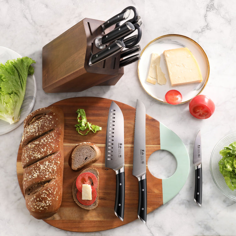 Wooden knife block with seven knives next to a cutting board holding sliced bread, tomato, and herbs, with cheese and lettuce